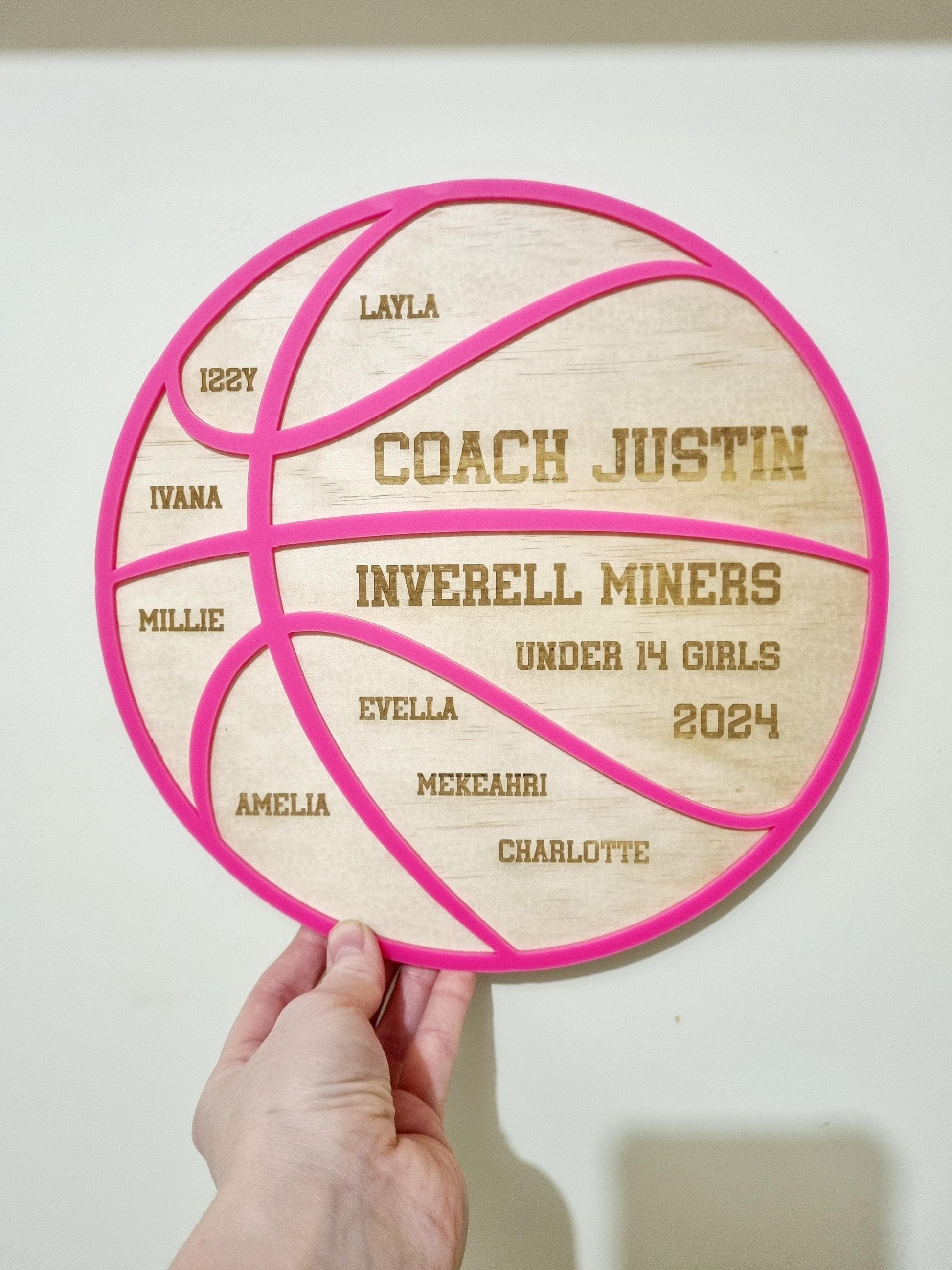 Personalised basketball coach gift set displayed on a white surface. The custom wooden plaque is shaped like a basketball with engraved team names, player name, and the coach’s name, accented with a hot pink acrylic overlay.