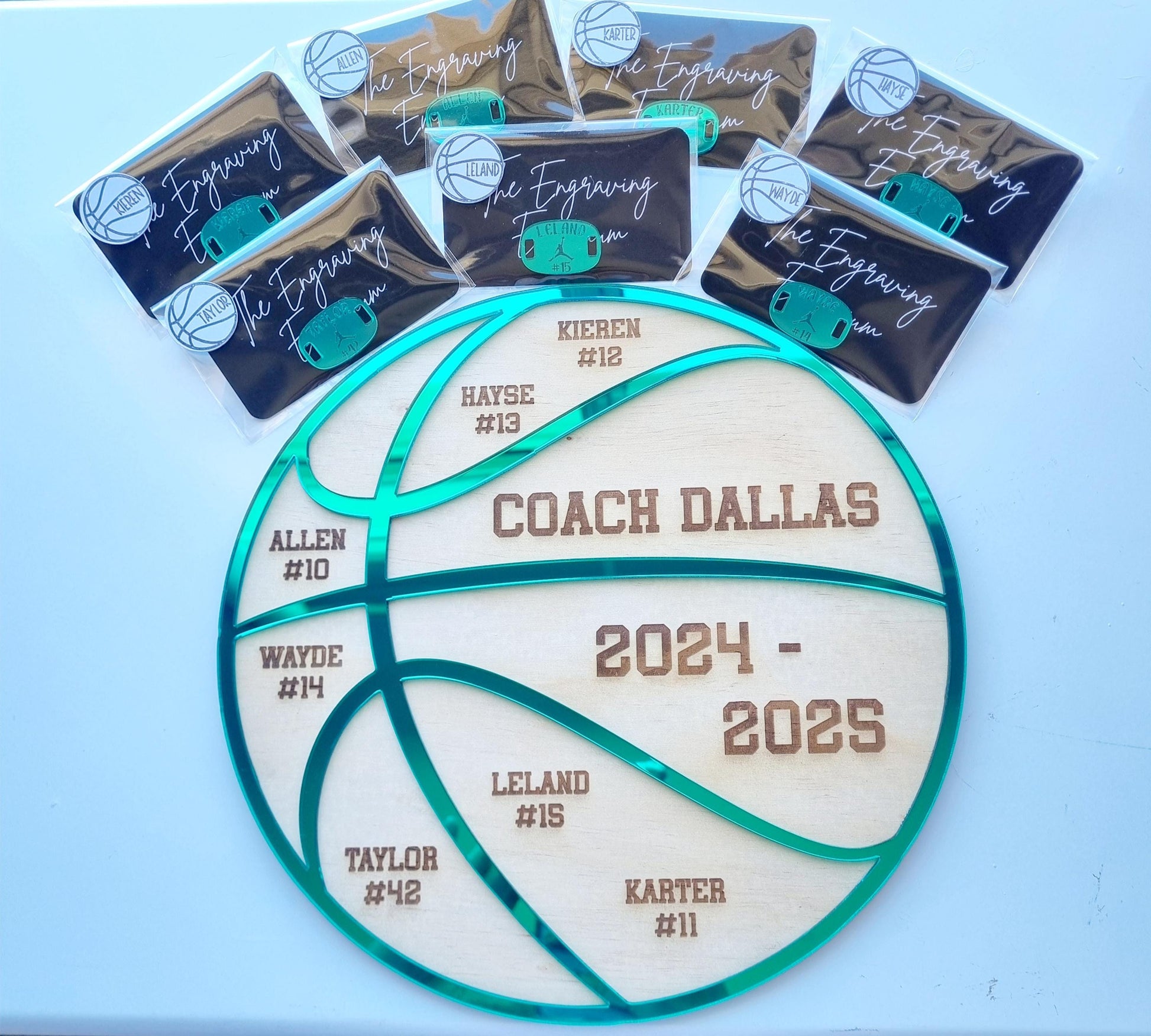 Personalised basketball coach gift set displayed on a white surface. The custom wooden plaque is shaped like a basketball with engraved team names, player numbers, and the coach’s name, accented with a green mirrored acrylic overlay. Surrounding the plaque are individually packaged engraved shoe tags, also made of green mirrored acrylic, featuring player names and numbers. A thoughtful and unique gift for basketball coaches.